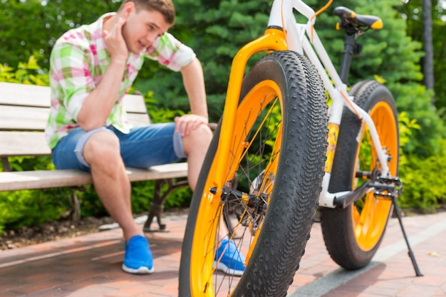 Hombre sentado con expresión triste sentado en un banco cerca de la bicicleta, se centran en la rueda en el hermoso parque verde