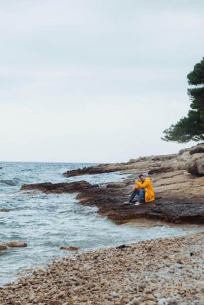 Hombre sentado en impermeable amarillo en la playa rocosa mirando el clima tormentoso