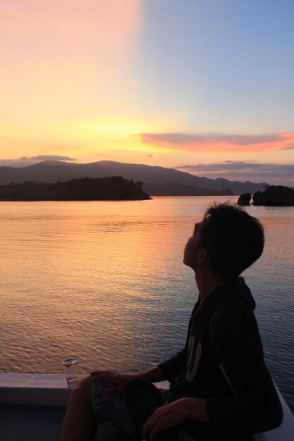 Hombre sentado junto al mar contra el cielo durante la puesta de sol