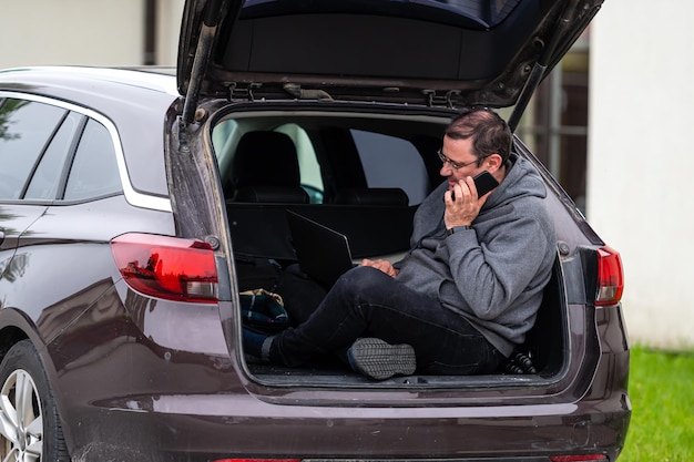 Un hombre sentado en el maletero abierto de un coche y trabajando con un trabajo remoto de tecnología móvil portátil