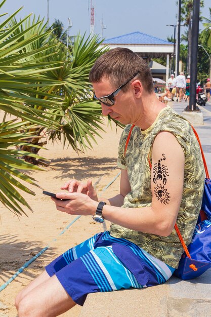 Hombre sentado en las manos de la playa sosteniendo un teléfono inteligente con un tatuaje de henna recién aplicado en el antebrazo