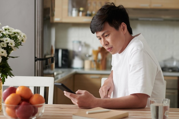 Hombre sentado en la mesa de la cocina con el ceño fruncido y leyendo notificaciones en el teléfono inteligente