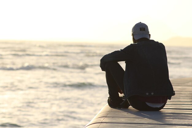 Foto hombre sentado en un muro de contención contra el mar durante la puesta de sol