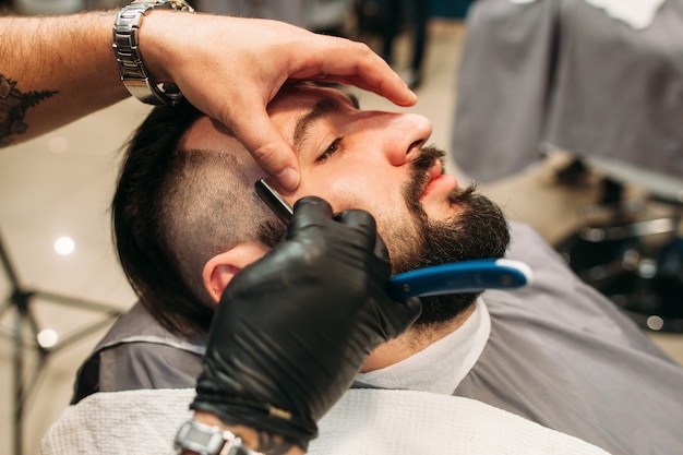 Foto hombre sentado con los ojos cerrados mientras el peluquero lo afeita con navaja