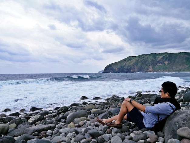 Foto hombre sentado en piedras de guijarros en la playa contra el cielo