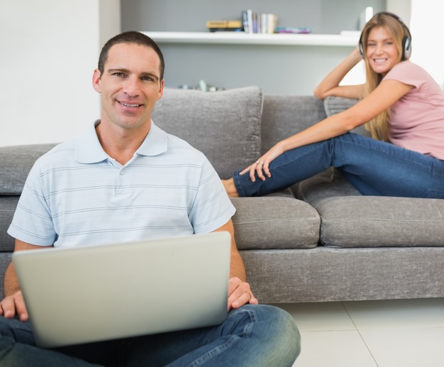 Hombre sentado en el piso con la computadora portátil con la mujer escuchando música en el sofá ambos mirando a la cámara