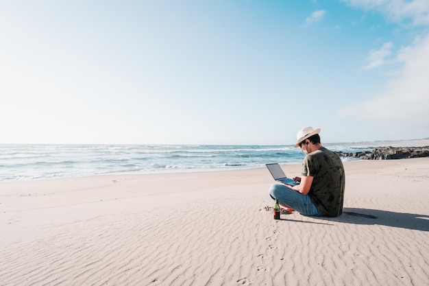 Hombre sentado en la playa bebiendo cerveza con un portátil solo