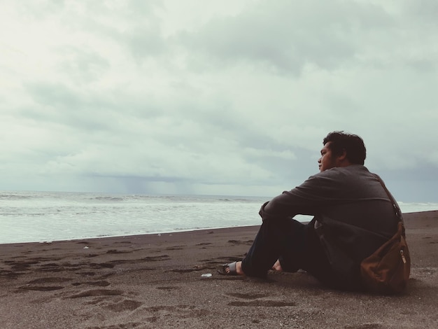 Foto hombre sentado en la playa mirando al mar contra el cielo