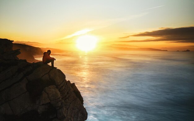 Foto hombre sentado en rocas junto al mar contra el cielo durante la puesta de sol