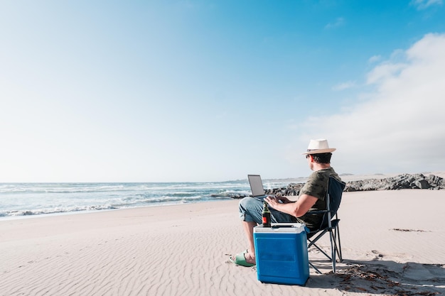 Hombre sentado en una silla de camping en la playa bebiendo cerveza con el portátil solo disfrutando