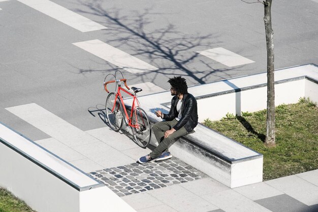 Hombre sentado en el skatepark de la ciudad sosteniendo su teléfono inteligente al lado de su bicicleta