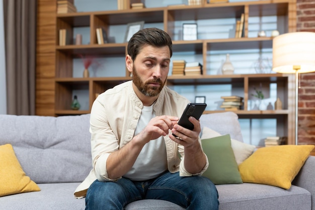 Un hombre sentado en un sofá en casa con aspecto aburrido mientras se desplaza por las aplicaciones de citas en su teléfono.