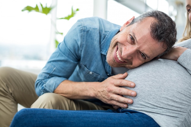 Hombre sentado en el sofá y escuchando el estómago de la mujer embarazada