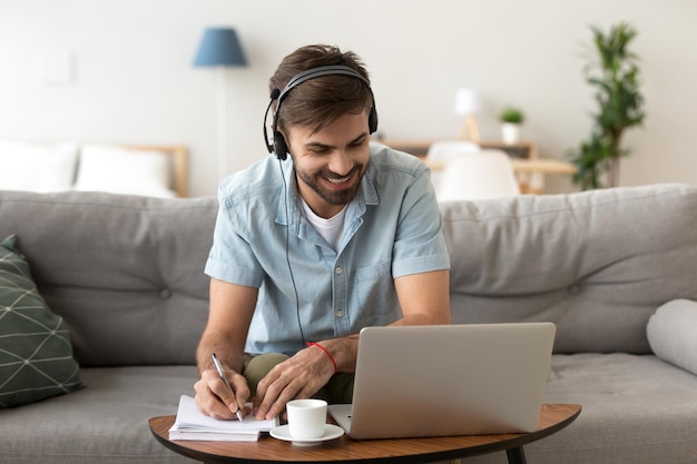 Hombre sentado en el sofá en la sala de estar en casa disfrutando de estudiar usando portátil y auriculares mirando la pantalla del dispositivo escuchando audio tomando algunas notas Hombre tiene lección en línea de aprendizaje electrónico en el concepto de internet