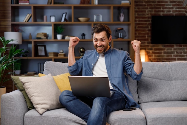 Hombre sentado en el sofá trabajando en una computadora portátil que recibe un correo electrónico de notificación de aprobación de préstamo bancario sintiéndose exitoso apretando los puños haciendo un gesto de sí Emociones sinceras victoria resultado logro concepto