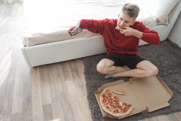 Hombre sentado en el suelo en una habitación con una caja de pizza y cambiar de canal de televisión