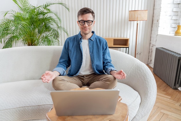 Foto un hombre sereno con gafas meditando frente a una computadora portátil en un sofá en un espacio de vida decorado con plantas