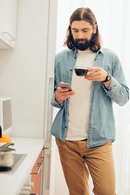 Hombre serio y atractivo que pasa tiempo en Internet en su teléfono inteligente y bebe café en casa