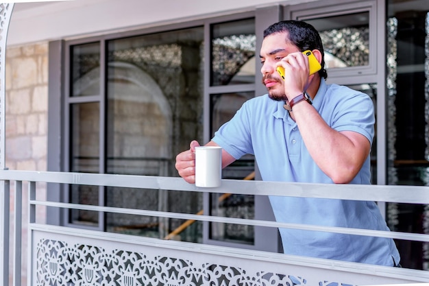 Hombre serio con barba hablando por teléfono inteligente con una taza de café en el balcón