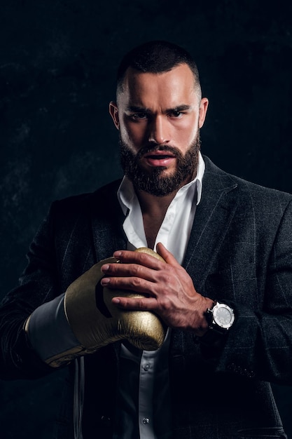 Un hombre serio y brutal con traje y guante de boxeo dorado está posando para un fotógrafo en un estudio fotográfico oscuro.