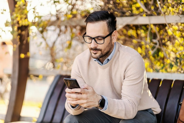 Un hombre serio en causal inteligente está sentado en un banco afuera y enviando mensajes de texto a su teléfono celular