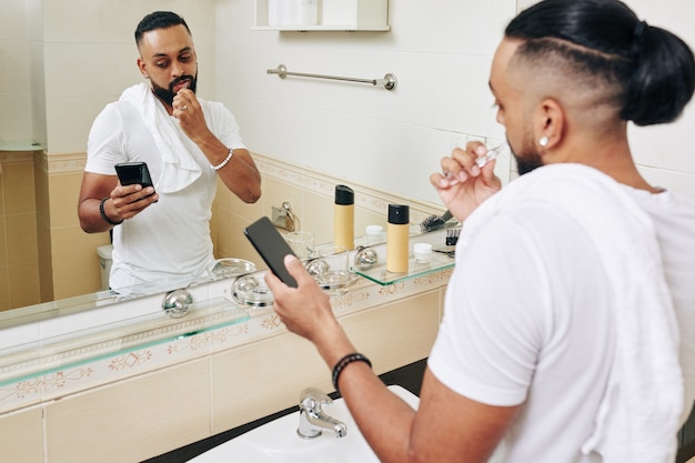 Hombre serio cepillarse los dientes en el baño y leer noticias en la pantalla del teléfono inteligente