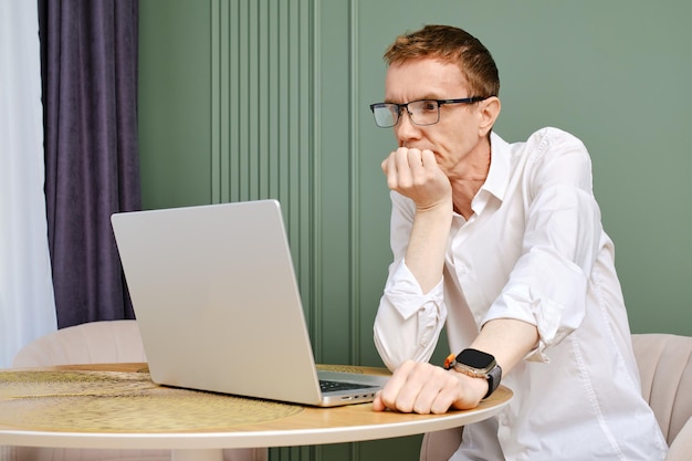 Hombre serio y concentrado de mediana edad sentado en casa frente a la computadora portátil