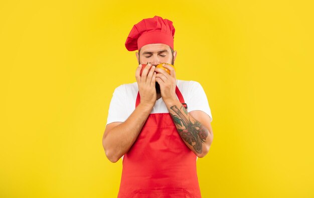 Hombre serio en delantal de cocina y toque oliendo tomates con los ojos cerrados chef de fondo amarillo