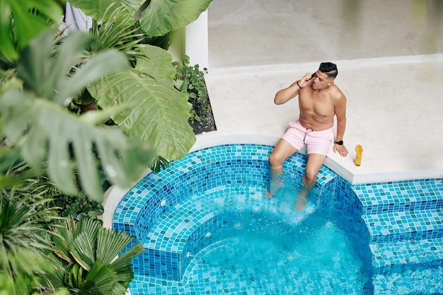 Hombre serio en forma tomando el sol en la piscina, bebiendo jugo de naranja y hablando por teléfono