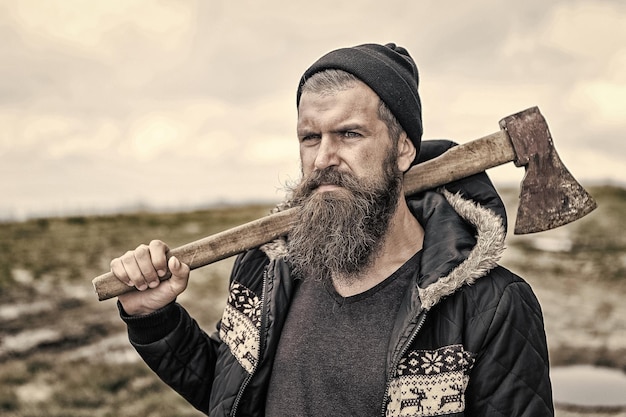 Hombre serio guapo barbudo con hacha oxidada en la cima de la montaña