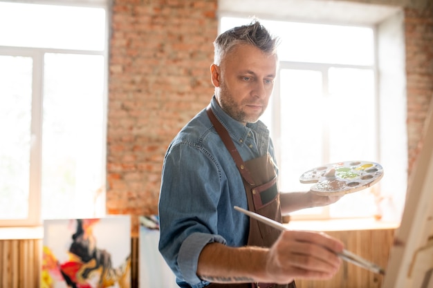 Foto hombre serio inspirado con paleta y pincel de pie delante de caballete y pintura en taller o estudio