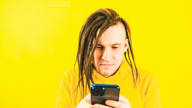 Hombre serio joven navegando por teléfono móvil sobre fondo amarillo Hombre guapo en sudadera con capucha amarilla usando teléfono inteligente