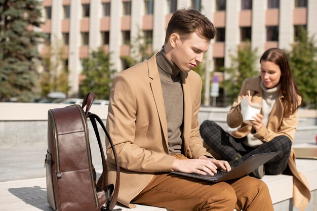 Hombre serio joven en ropa casual elegante usando la computadora portátil mientras está sentado en un banco en el entorno urbano en el fondo de su novia comiendo wok