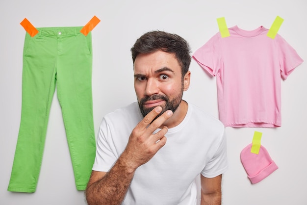 El hombre serio mantiene la mano en la barbilla, mira atentamente a la cámara, parece escrupuloso, levanta las cejas y examina algo, usa una camiseta casual aislada sobre la ropa de fondo blanco pegada detrás