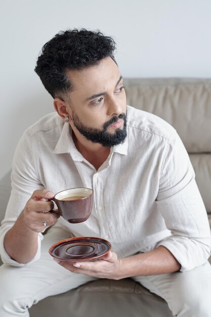 Hombre serio pensativo en loungewear sentado en el sofá bebiendo una taza de café y mirando a otro lado