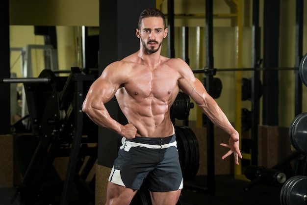 Hombre serio de pie fuerte en el gimnasio y flexionando los músculos