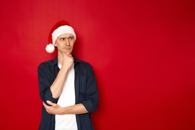 Hombre serio en pose pensativa sostiene la mano debajo de la barbilla en sombrero de navidad aislado sobre fondo rojo.