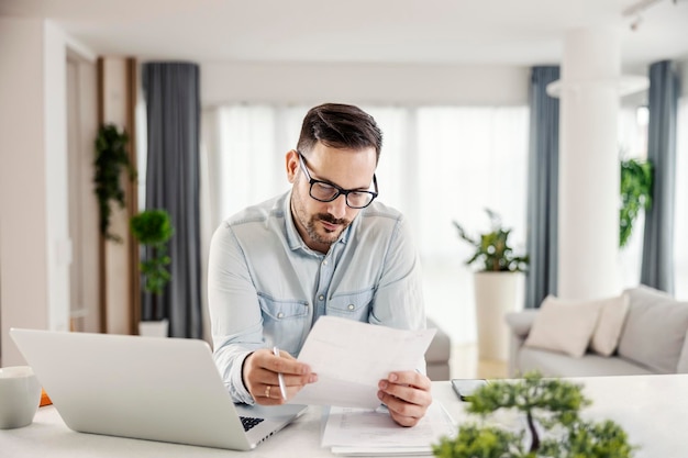 Un hombre serio que mira el papeleo y las facturas mientras está de pie junto a una computadora portátil en la oficina de su casa