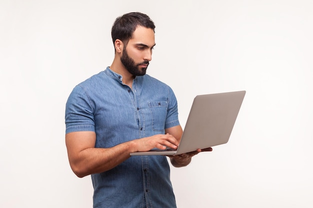 Hombre serio seguro de sí mismo con barba en camisa azul sosteniendo una computadora inalámbrica en la mano trabajando en una computadora portátil haciendo ediciones límite Disparo de estudio interior aislado sobre fondo blanco