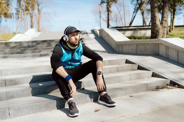 Un hombre serio se sienta en los escalones del parque con los brazos cruzados escucha música con auriculares inalámbricos