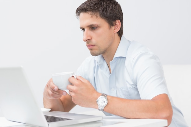 Hombre serio con taza de té usando la computadora portátil en casa