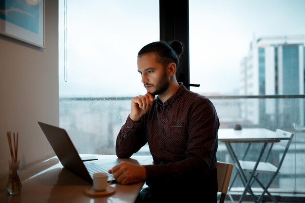 Hombre serio trabajando en casa en una computadora portátil Con fondo de ventanas panorámicas