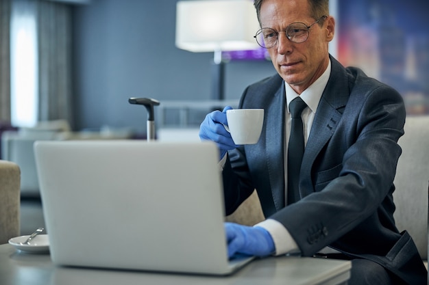 Hombre serio con traje y gafas tomando café y usando un portátil antes del vuelo durante la pandemia