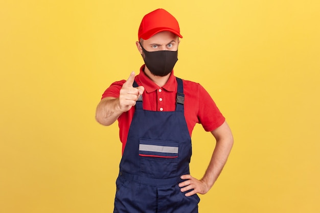 Hombre serio en uniforme de trabajador y máscara protectora mostrando el dedo mirando a la cámara