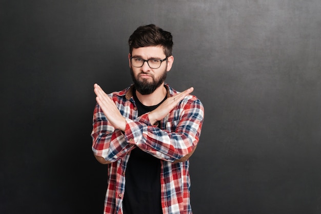 Hombre serio vestido con camisa en una jaula y con gafas de pie en la pizarra mientras hace el gesto de parada