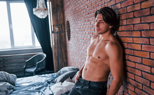 Hombre sexy sin camisa apoyado en la pared de ladrillo en el dormitorio a la hora de la mañana.