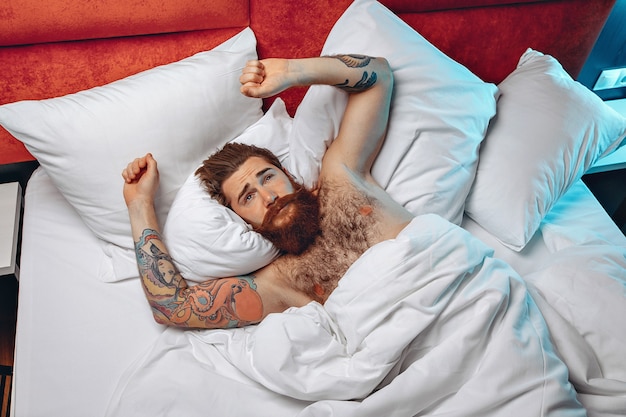 Hombre sexy varonil con un largo bigote, barba y tatuajes que acaba de despertar y estirar en su cama blanca, abriendo los ojos temprano en la mañana.