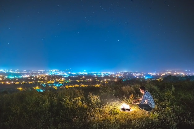 El hombre se sienta cerca de la hoguera en el fondo de las luces de la ciudad. Noche