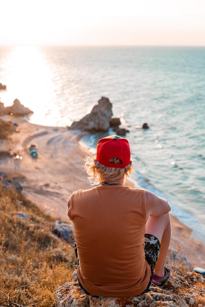 Foto hombre se sienta en la cima de una montaña en la orilla del mar y mira la puesta de sol viajes y turismo