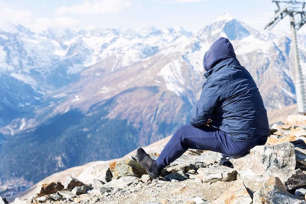 Un hombre se sienta en lo alto de las montañas y admira el paisaje. Un turista con una chaqueta con capucha azul en una naturaleza magnífica. Turismo activo y deportes de invierno.